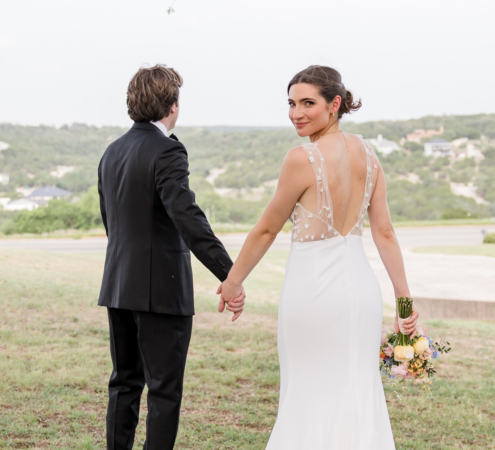 Back view of a newlywed couple holding hands and walking on a grassy hill overlooking a suburban valley. The bride is looking over her shoulder, showcasing her low-back gown with sheer lace appliqué and a long train.
