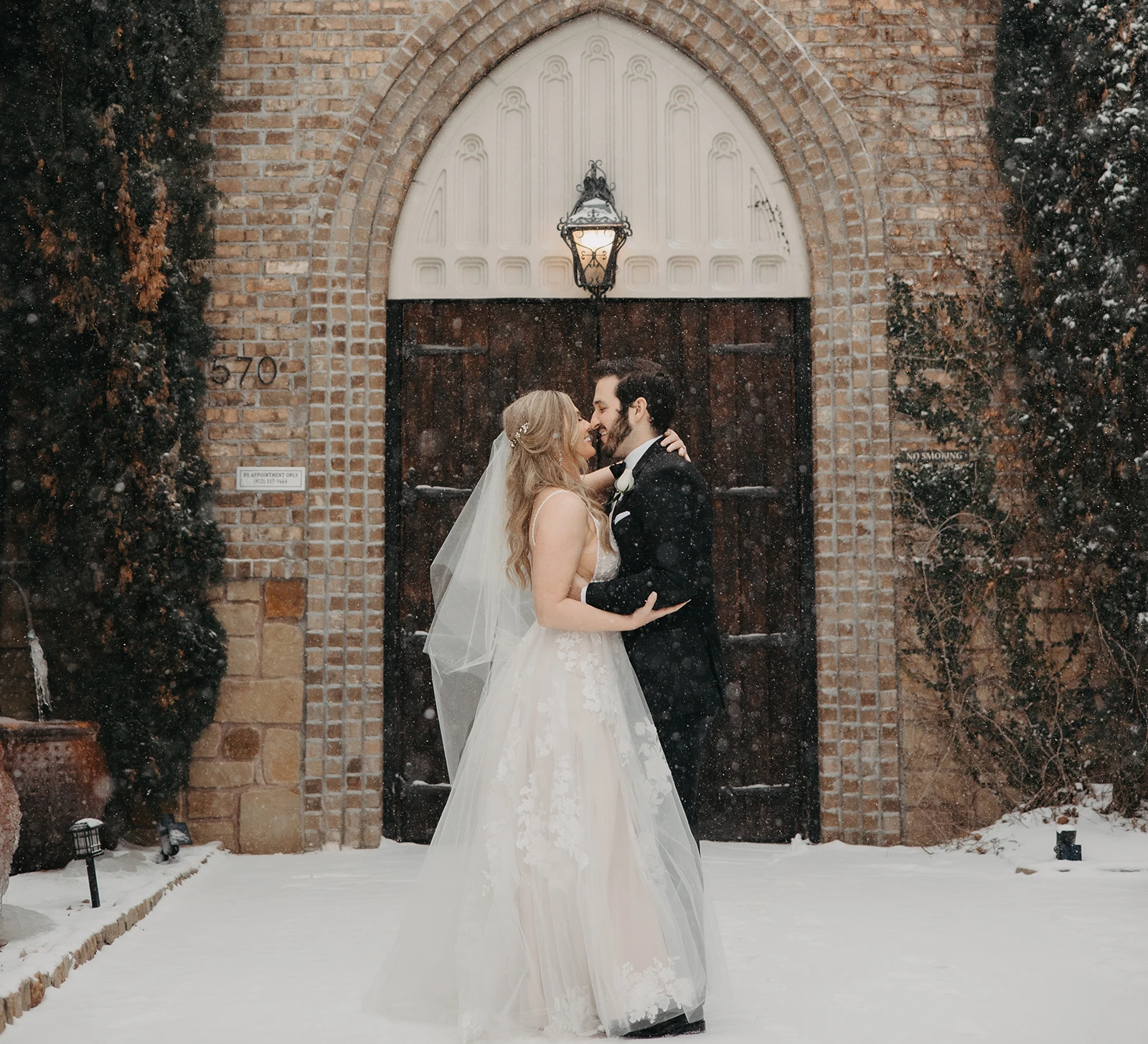 Romantic winter wedding portrait of the bride and groom kissing in falling snow outside a brick chapel with a terracotta tile roof. The bride wears a sheer lace gown, and the ground is covered in white snow.