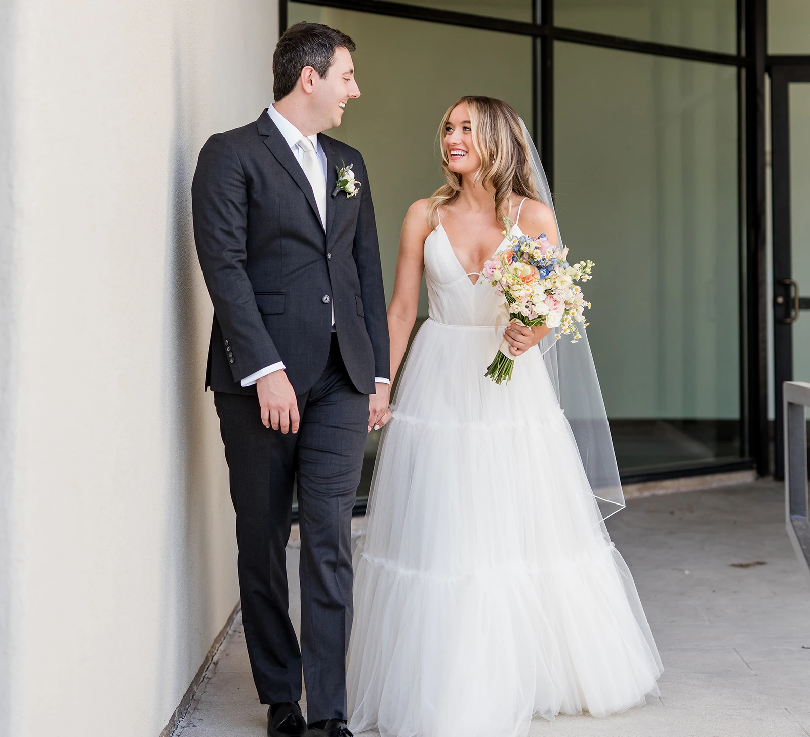 Bride and groom smiling and holding hands during their first look, with the bride in a tulle gown holding a pastel bouquet