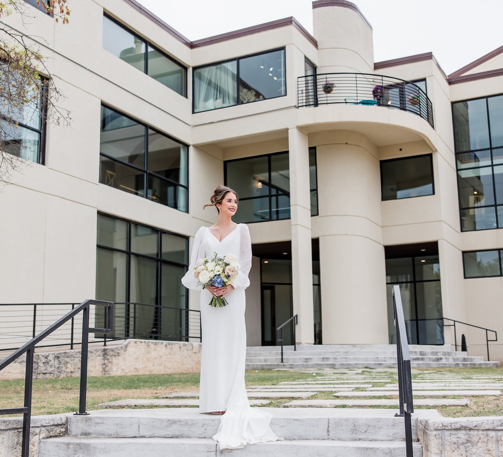 Bridal portrait of the bride standing on concrete steps in front of a modern, architectural wedding venue with sweeping lines and black-framed windows. The bride wears a sleek white gown with sheer, voluminous sleeves and holds a mixed floral bouquet.