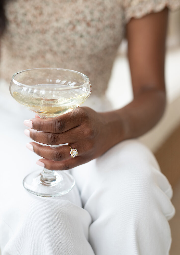 A flat lay of gold wedding jewelry, including rings and bracelets, displayed on a dark green velvet surface next to monogrammed gift boxes and champagne glasses.