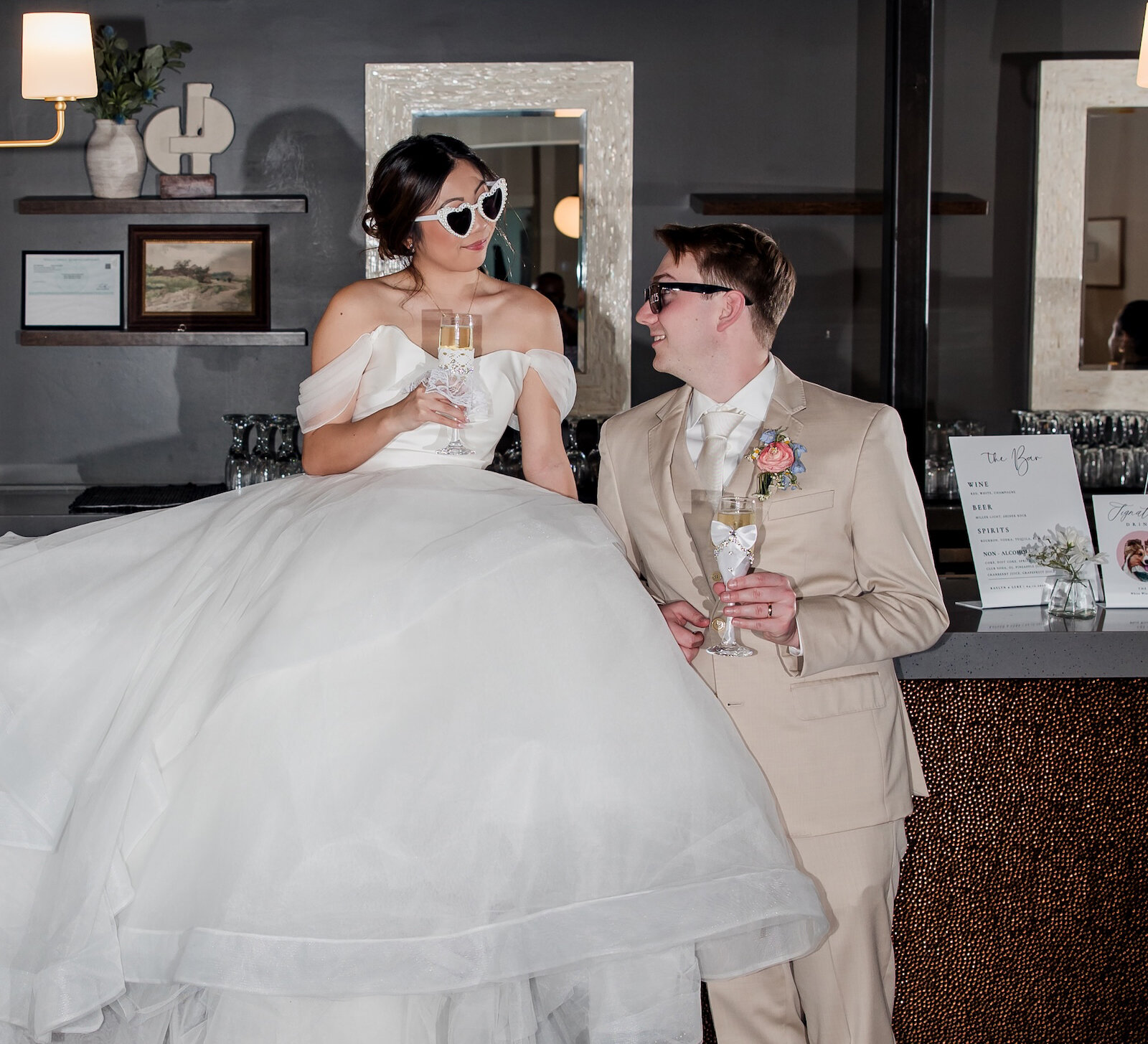 Fun and stylish portrait of the newlyweds at the reception bar. The bride is seated on the bar in a full white tulle ballgown and heart-shaped sunglasses, sharing a champagne toast with the groom, who is wearing a tan suit.