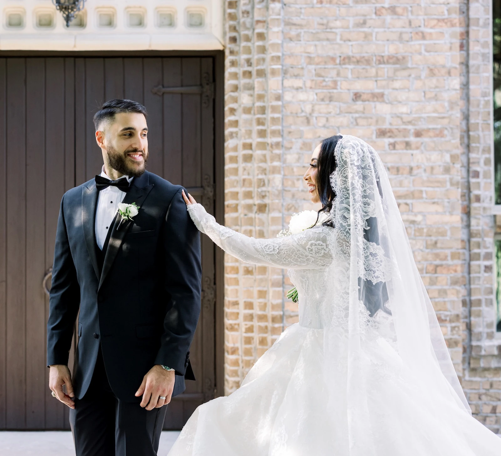 Elegant wedding portrait of the bride and groom smiling outside a brick building with dark wooden doors. The groom wears a black tuxedo, and the bride wears a long-sleeved lace gown with a dramatic, long lace-edged Spanish veil.