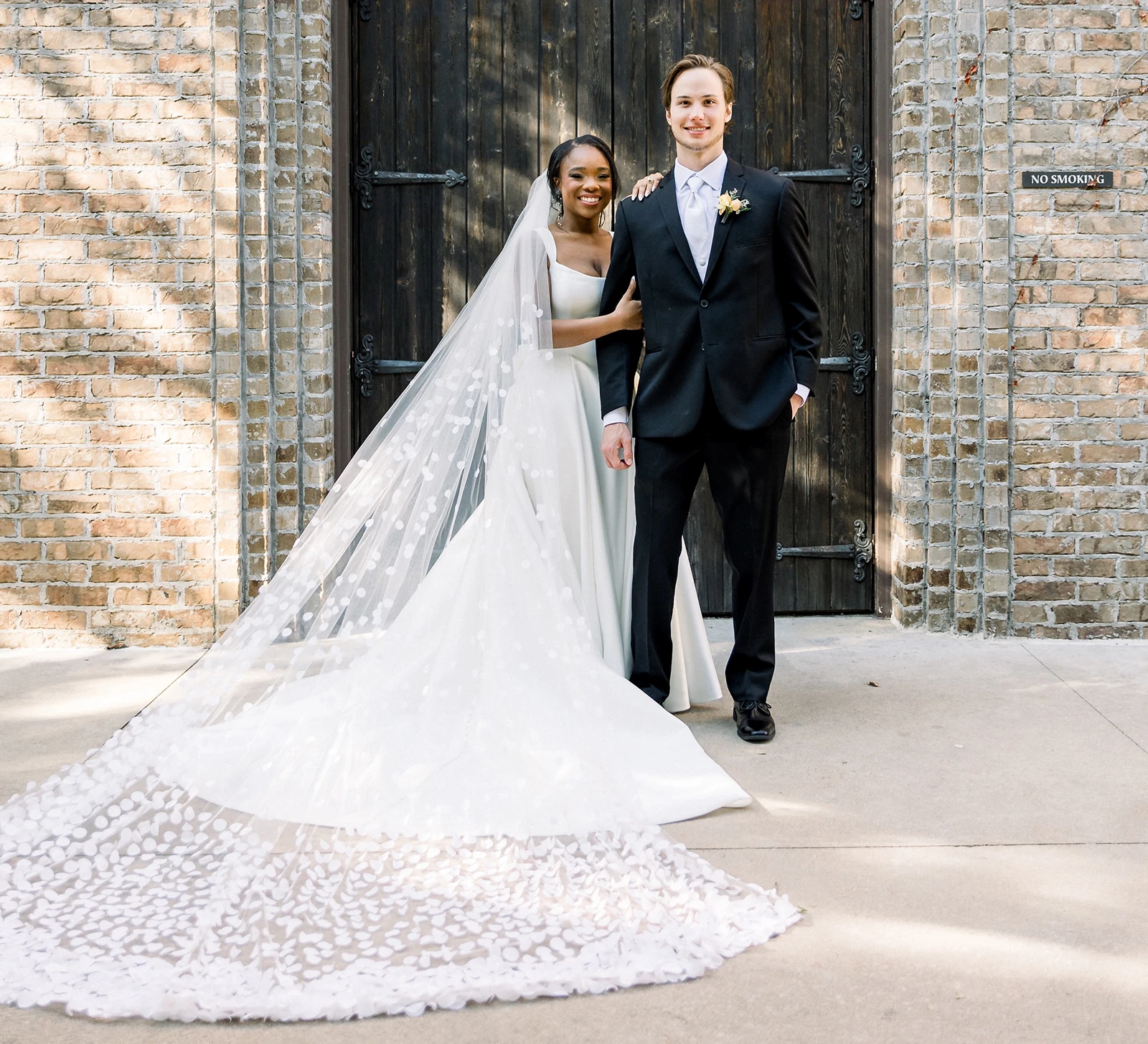 Elegant portrait of the wedding couple standing in front of a Gothic arched entryway with dark wooden doors and a brick facade. The bride wears a sleek white gown with a dramatic cathedral-length lace veil spread out on the ground.