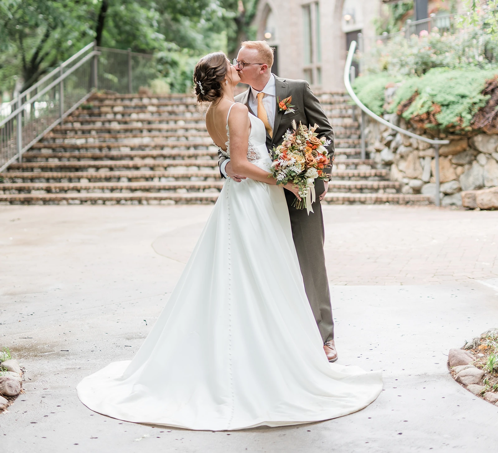 Bride and groom share a kiss outdoors in front of stone steps, with the bride’s long white gown flowing behind her and the groom holding an autumn-colored bouquet