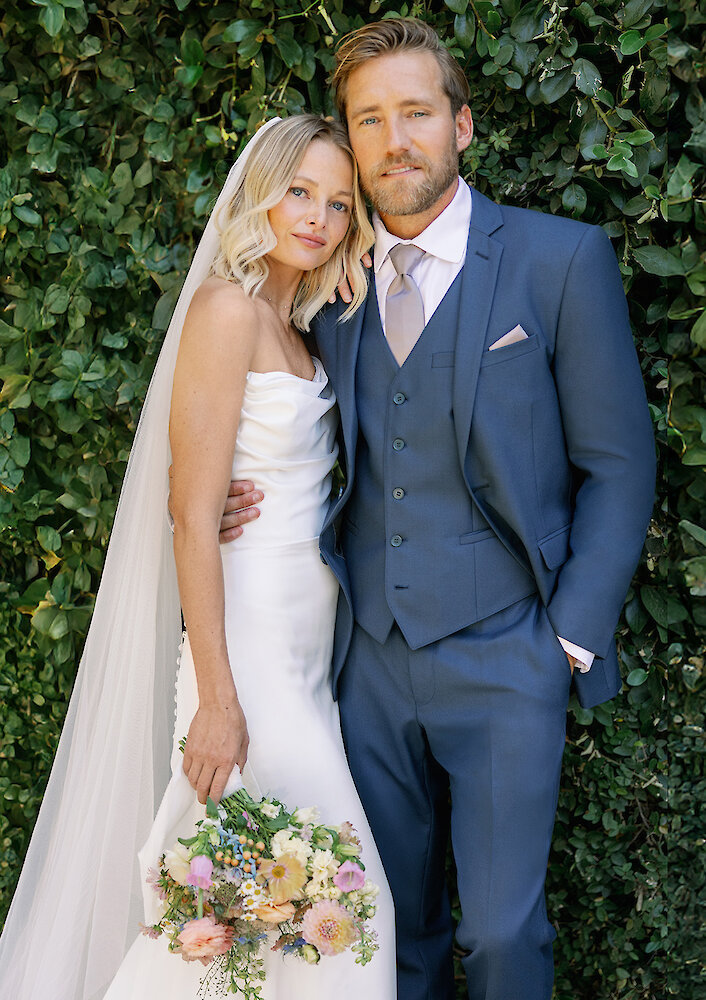 Fun and unconventional wedding portrait of the bride and groom sitting relaxed on a leather armchair, eating slices of pizza. The bride is barefoot in her voluminous ballgown, and the groom wears a black tuxedo in a formal, dark blue paneled room.