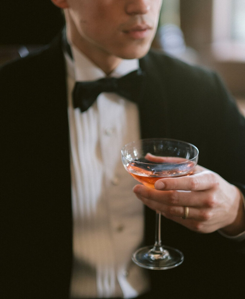 A close-up of a groom wearing a tuxedo and black bow tie, holding a coupe glass filled with a pink cocktail.
