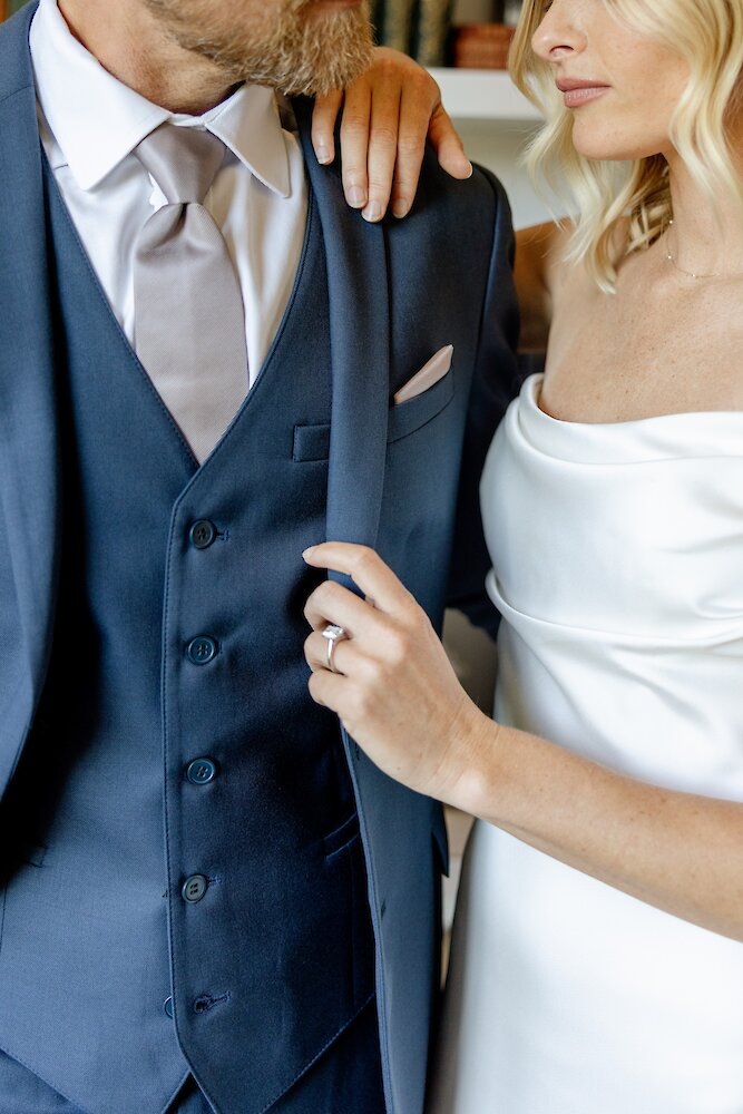 A close-up of the bride and groom, showing the groom's navy three-piece suit and the bride's hand with an engagement ring resting on his lapel.