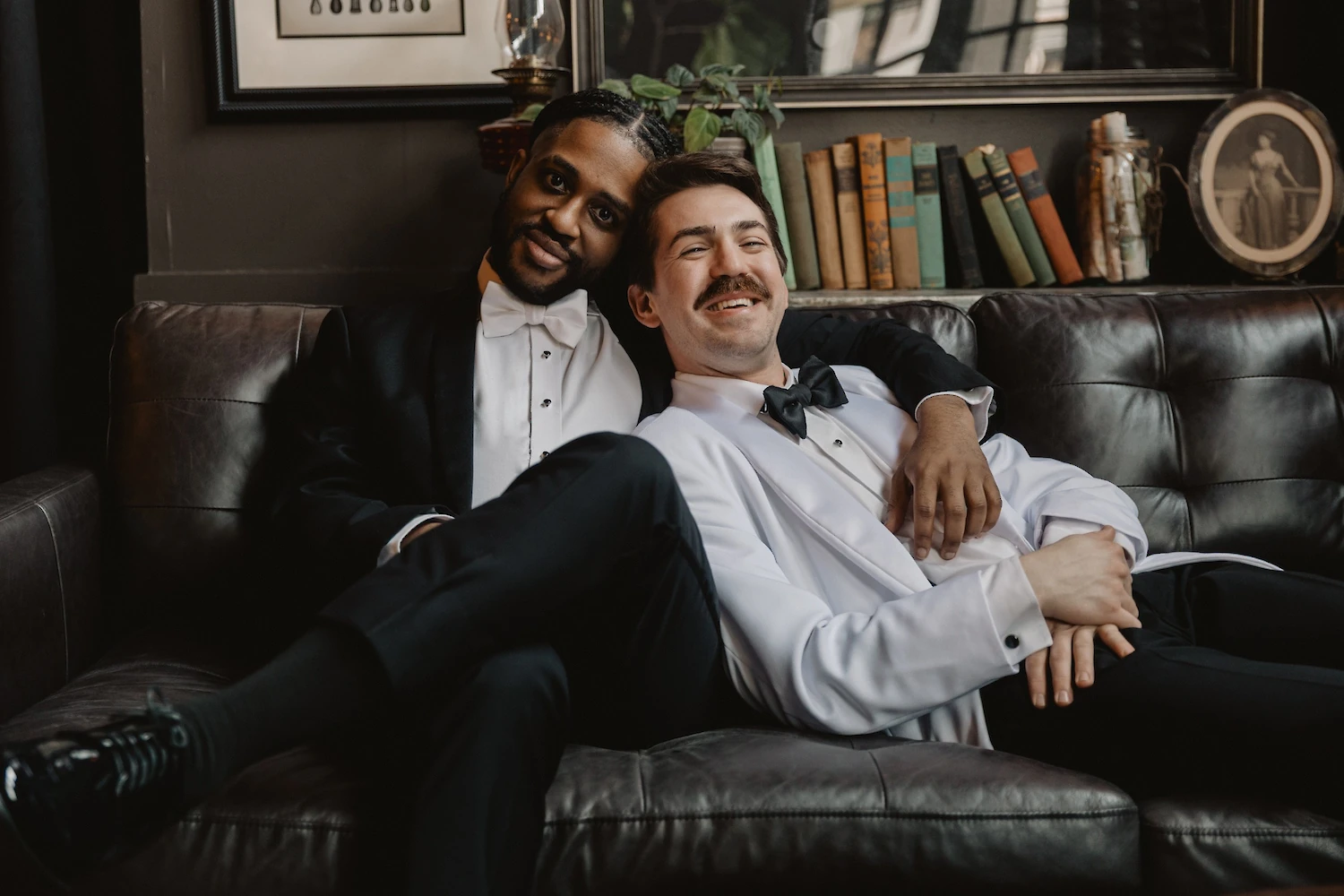 Two grooms, one in a black tuxedo and the other in a white jacket, are affectionately lounging together on a dark leather couch in a dimly lit room.