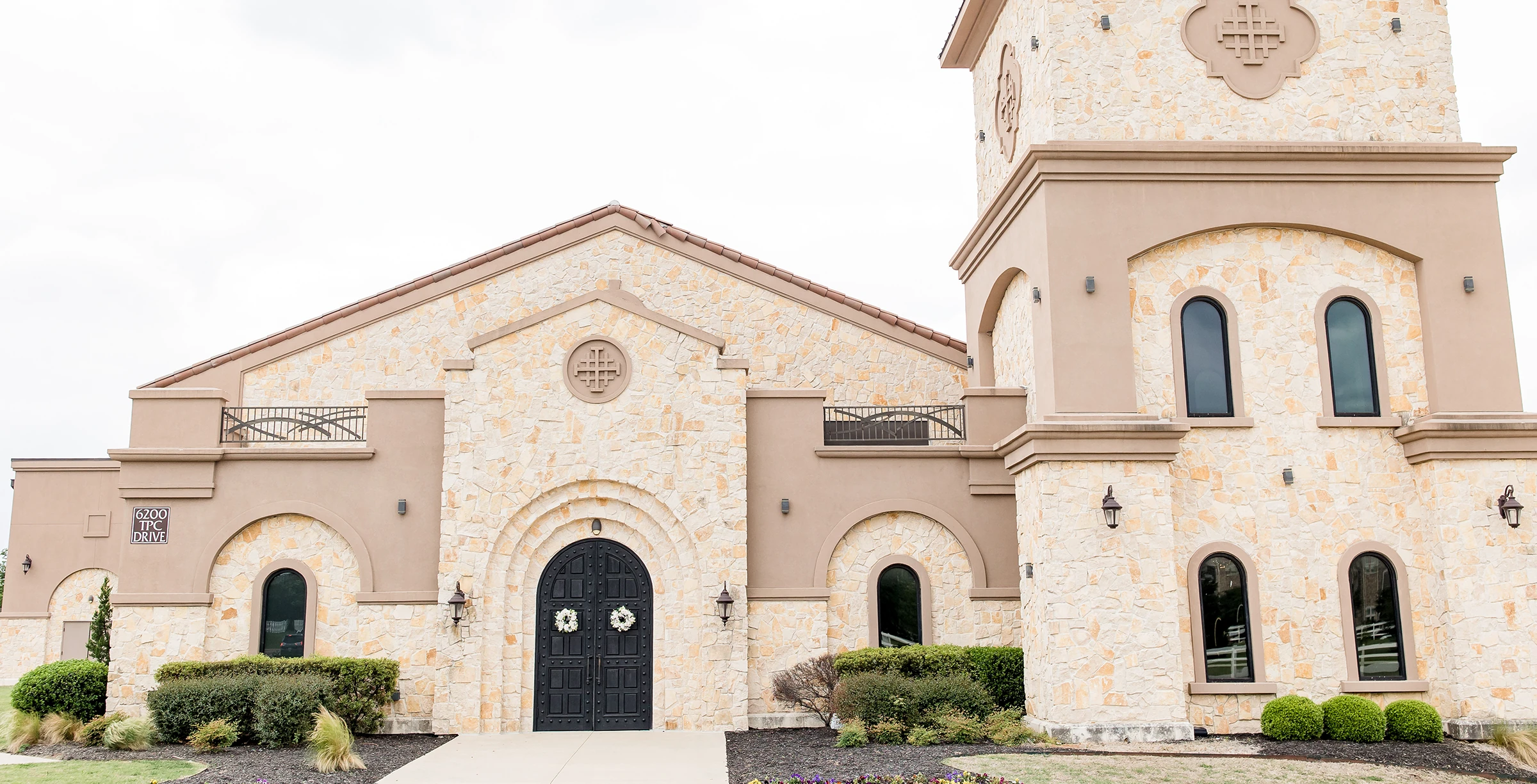 Exterior of a Mediterranean-style wedding venue with light stone walls, arched black double doors, and tall arched windows.