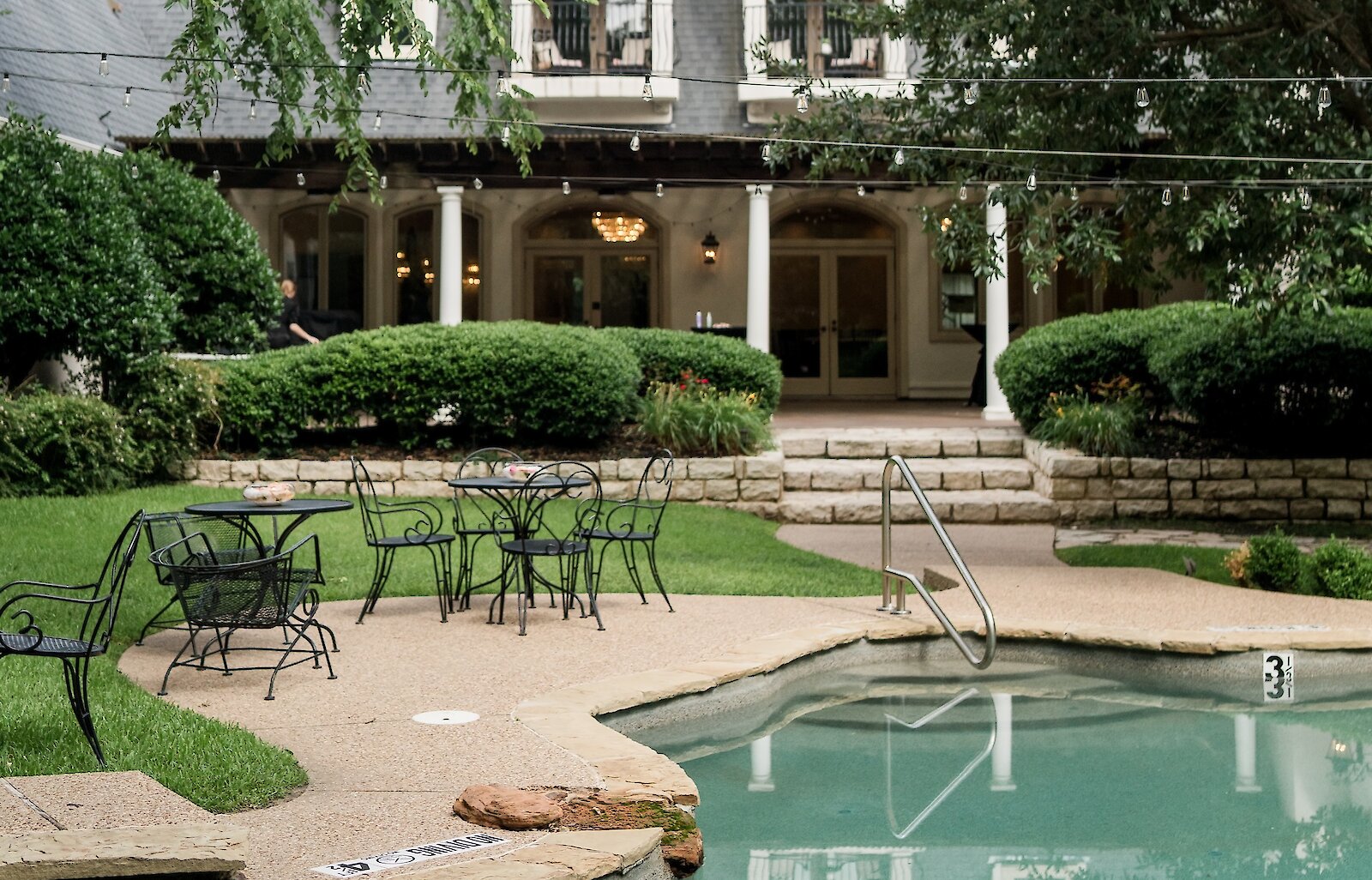 The exterior of a mansion venue featuring a backyard area with a swimming pool, stone patio, black wrought iron tables, and surrounding greenery.