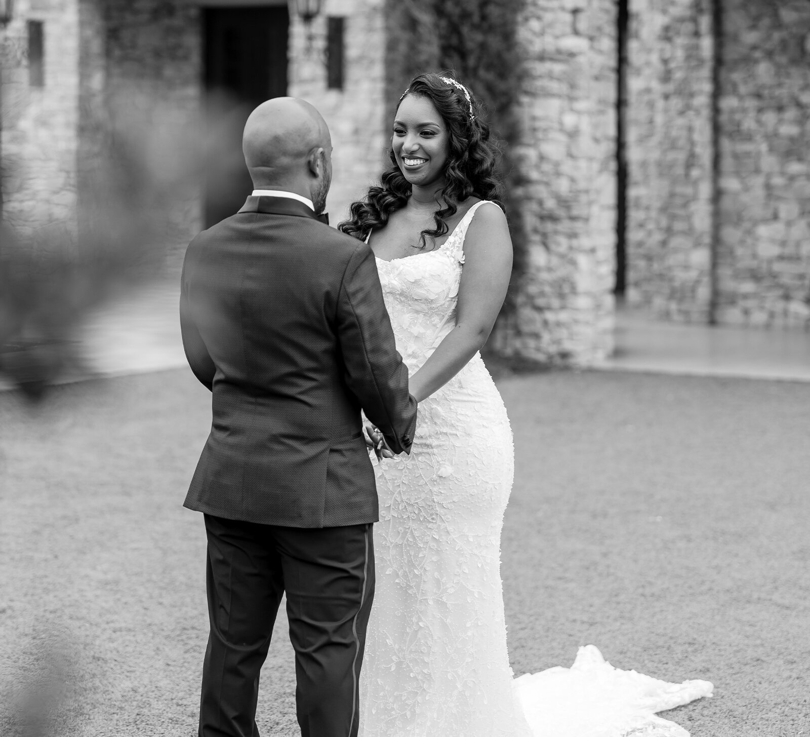 Close-up of a bride and groom reaching toward each other, showing their wedding rings with a blurred outdoor fountain in the background.