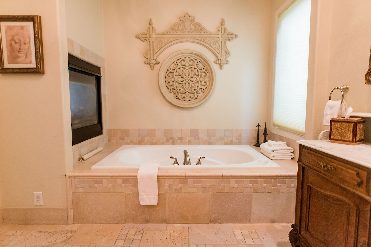 Elegant bathroom with a built-in soaking tub, beige tile surround, ornate wall décor, folded white towels, and a classic wooden vanity.