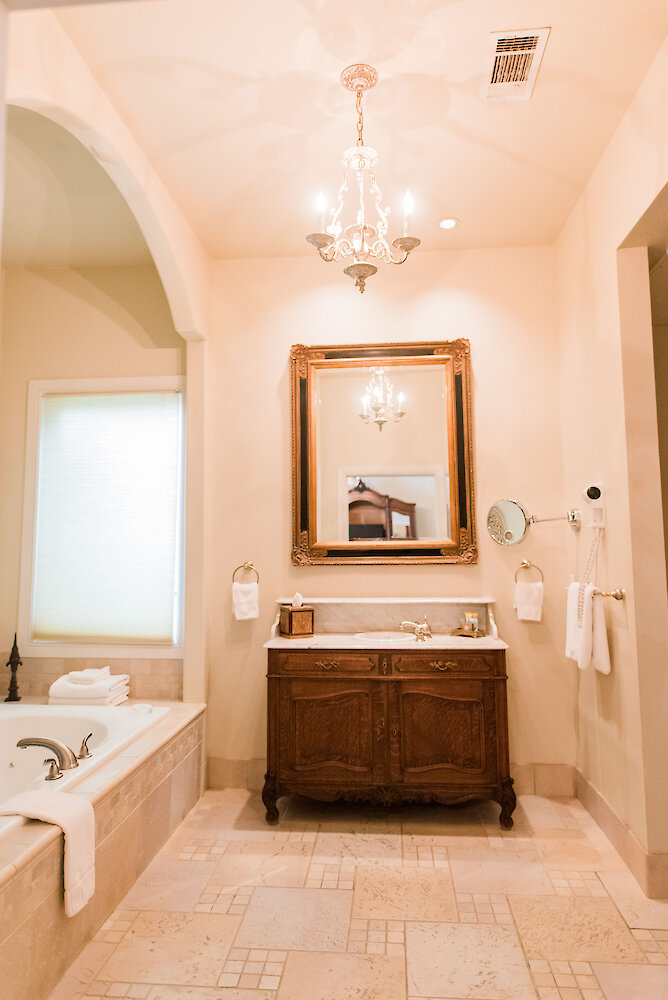 Elegant bathroom with a large framed mirror, ornate chandelier, wooden vanity with marble countertop, soaking tub, and soft neutral tile flooring