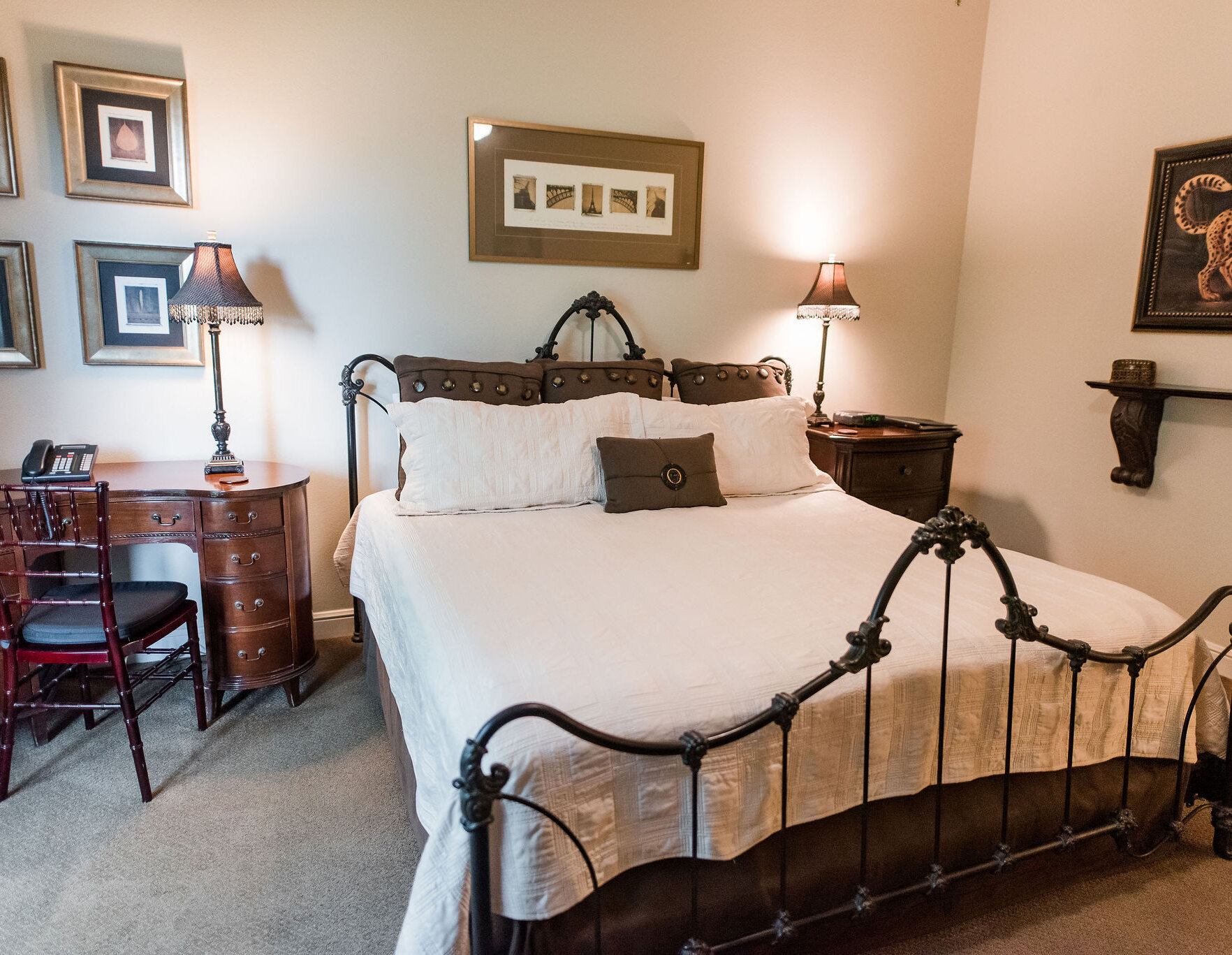 Cozy bedroom with an ornate metal bed frame, white bedding, wooden nightstands with decorative lamps, framed artwork, and a vintage wooden desk with chair.