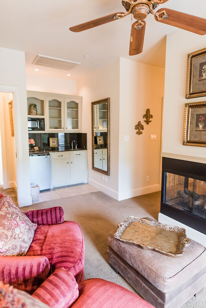 Cozy sitting area with red upholstered armchairs, an ottoman with a decorative tray, built-in kitchenette with glass cabinets, large wall mirror, and a double-sided fireplace