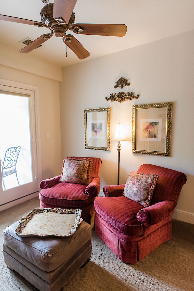 Cozy seating area with two red upholstered armchairs, floral accent pillows, a leather ottoman with a decorative tray, wall art in gold frames, and a floor lamp beside a glass door leading to a patio.