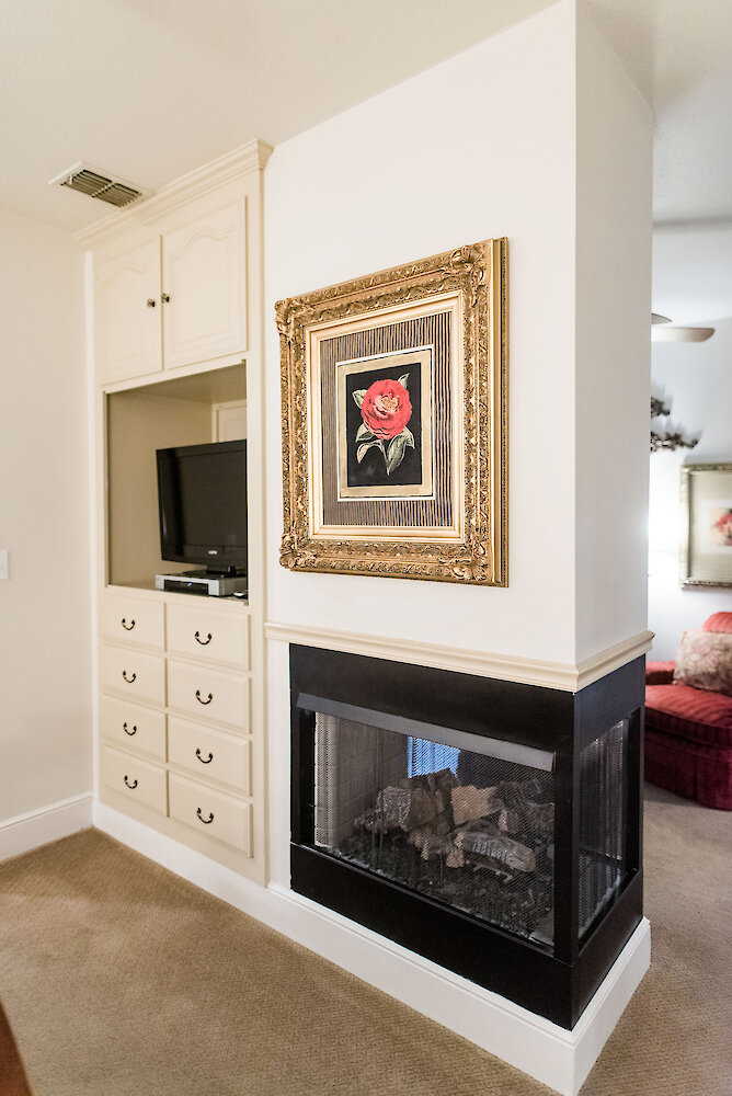 Interior view of a cozy suite featuring a built-in cabinet with a TV, a three-sided fireplace, and a framed red rose artwork in an ornate gold frame.