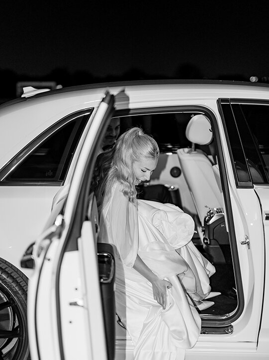 Bride stepping out of a white luxury car at night, holding her wedding dress as she exits, captured in black and white.