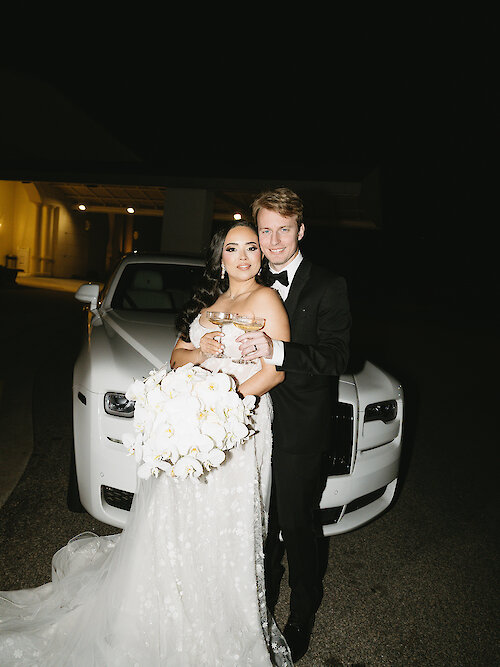 Bride and groom posing at night in front of a white luxury car, holding champagne glasses and a large white orchid bouquet.
