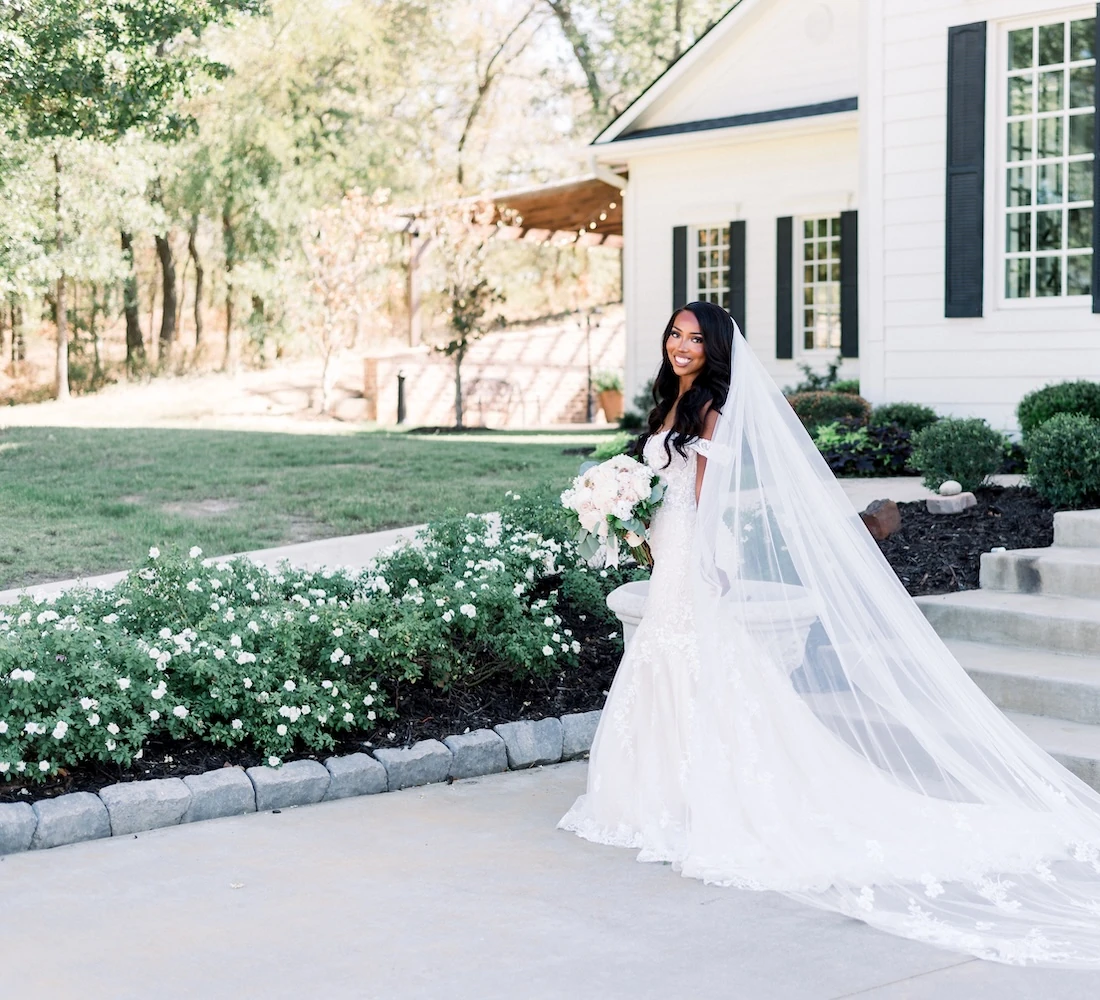Bride in a long lace wedding gown with a flowing veil holding a bouquet while standing outside a white Southern-style venue.