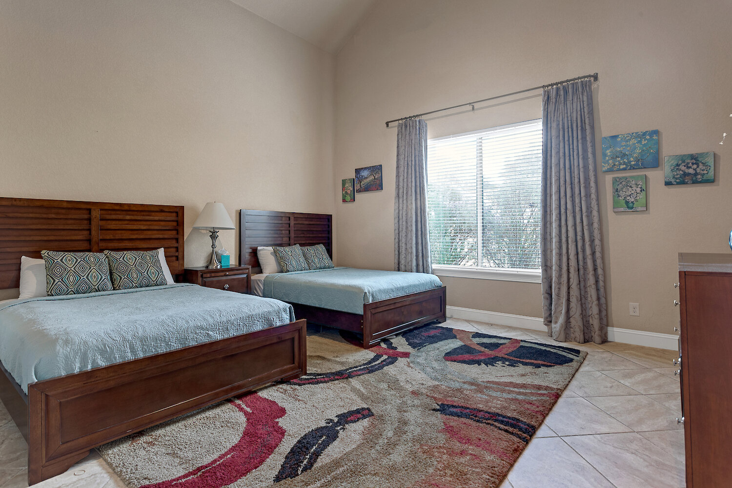 Spacious bedroom with two wooden beds, matching blue bedding, large window with curtains, and decorative wall art
