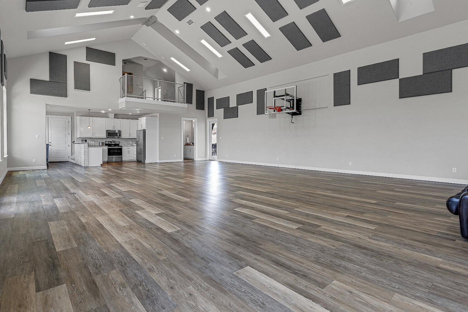 Large indoor multipurpose room with wood-look flooring, high ceilings with acoustic panels, basketball hoop, and open kitchen area.