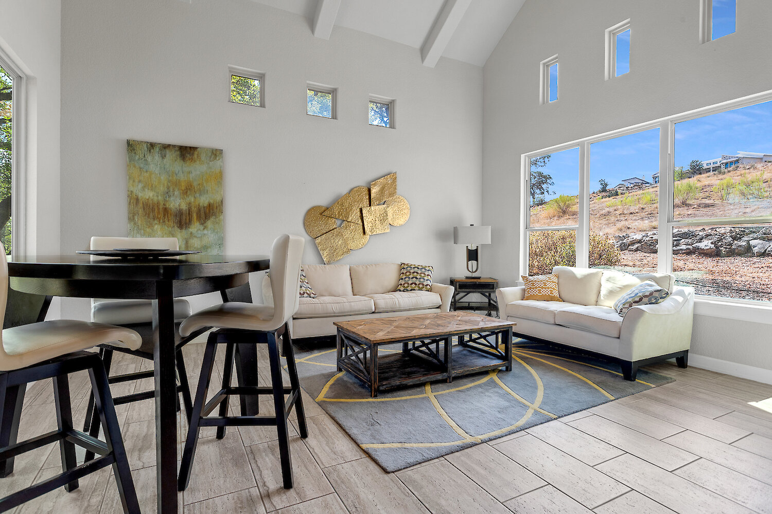 Bright modern living room with high ceilings, large windows, two cream sofas, abstract gold wall art, a round dining table, and a wooden coffee table on a patterned rug