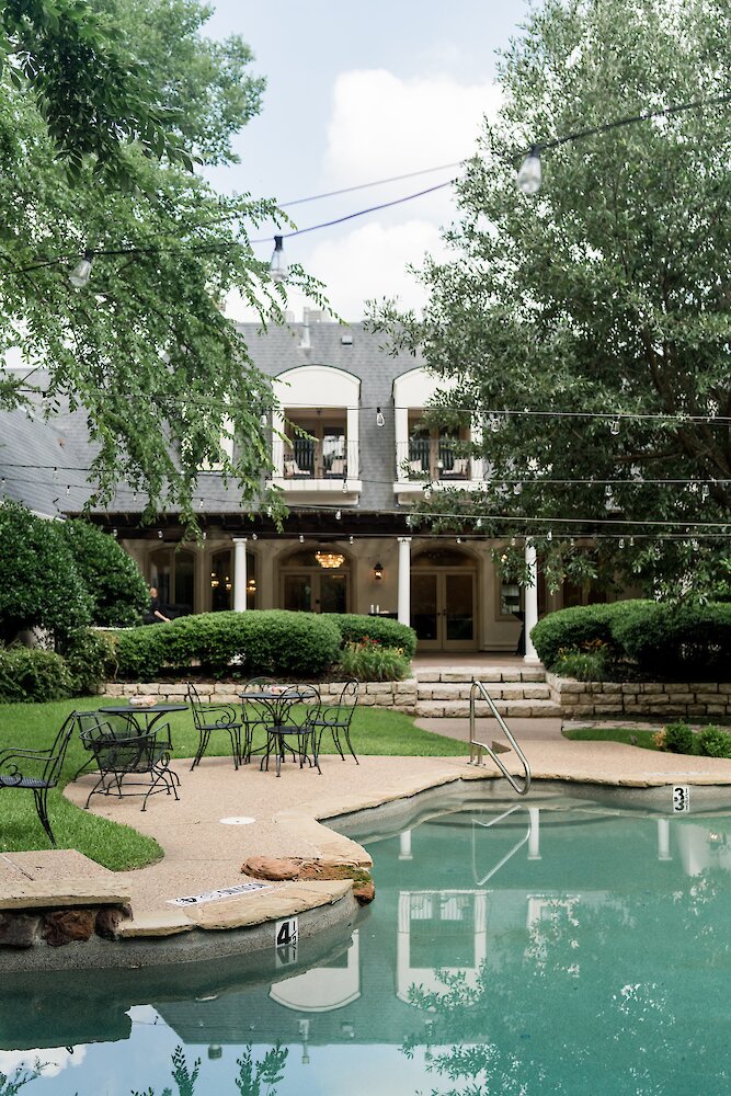 The exterior of a wedding mansion with dormer windows and a covered porch, overlooking a swimming pool and patio with wrought iron tables.