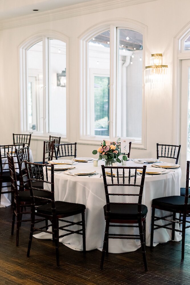 A wedding reception table with a white linen, gold charger plates, and a small floral centerpiece, set against a wall of arched white windows.