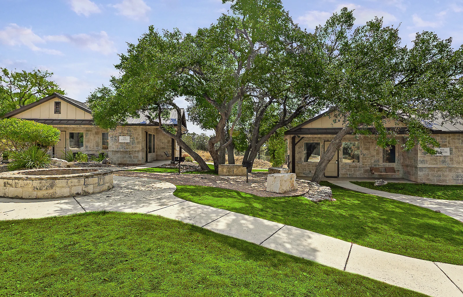 The exterior view of two small stone cottages with dark roofs, a winding concrete path, and a large central tree on a green lawn.