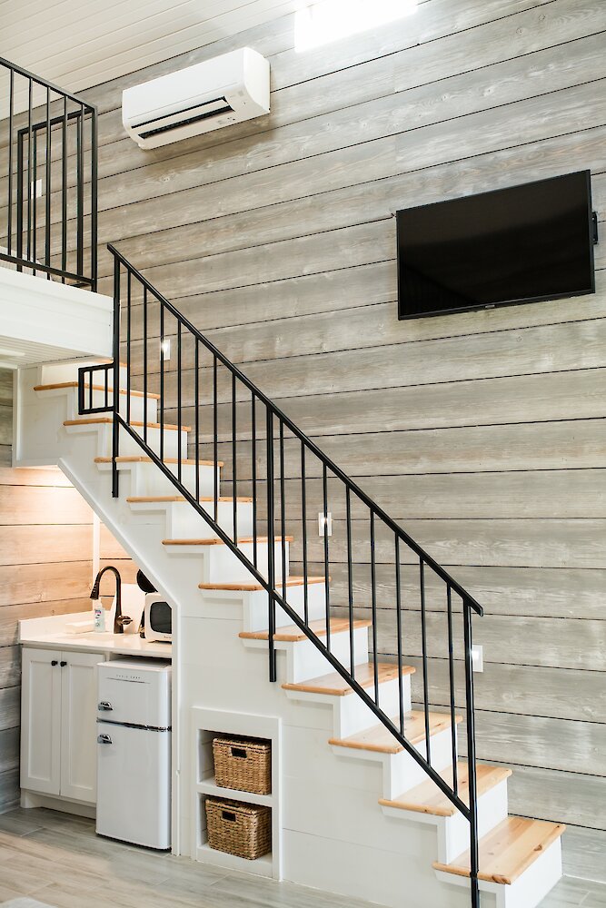 The interior of a modern cottage showing a white staircase with a black railing leading to a loft, with a small kitchenette built underneath.