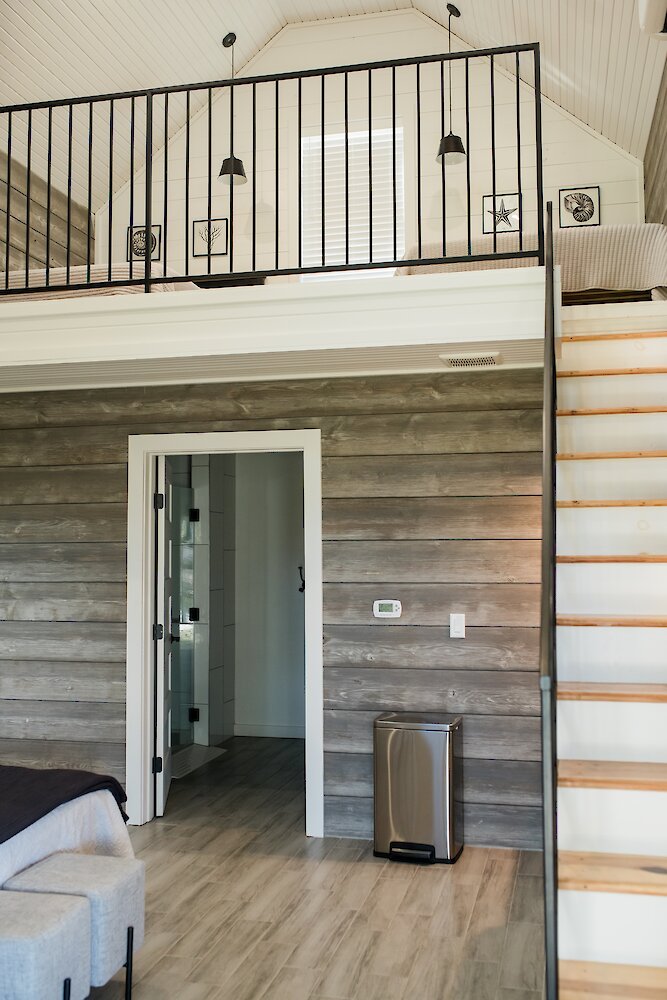The rustic interior of a cottage room featuring weathered wood plank walls, an open doorway, and a loft space accessed by a staircase with a black railing.