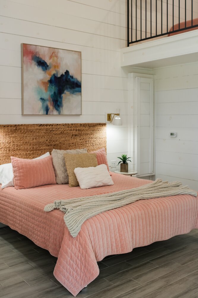 A cozy bedroom featuring a bed with a wicker headboard, a quilted pink velvet blanket, and abstract wall art above the headboard.