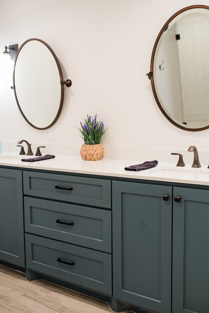 A modern bathroom double vanity with a dark teal cabinet base, oval framed mirrors, bronze faucets, and a small woven lavender plant.