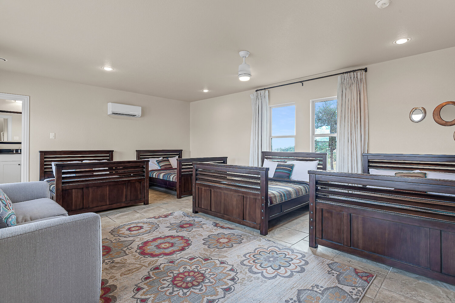 Spacious bunk-style bedroom with five wooden twin beds, patterned rug, tile flooring, and large windows with natural light