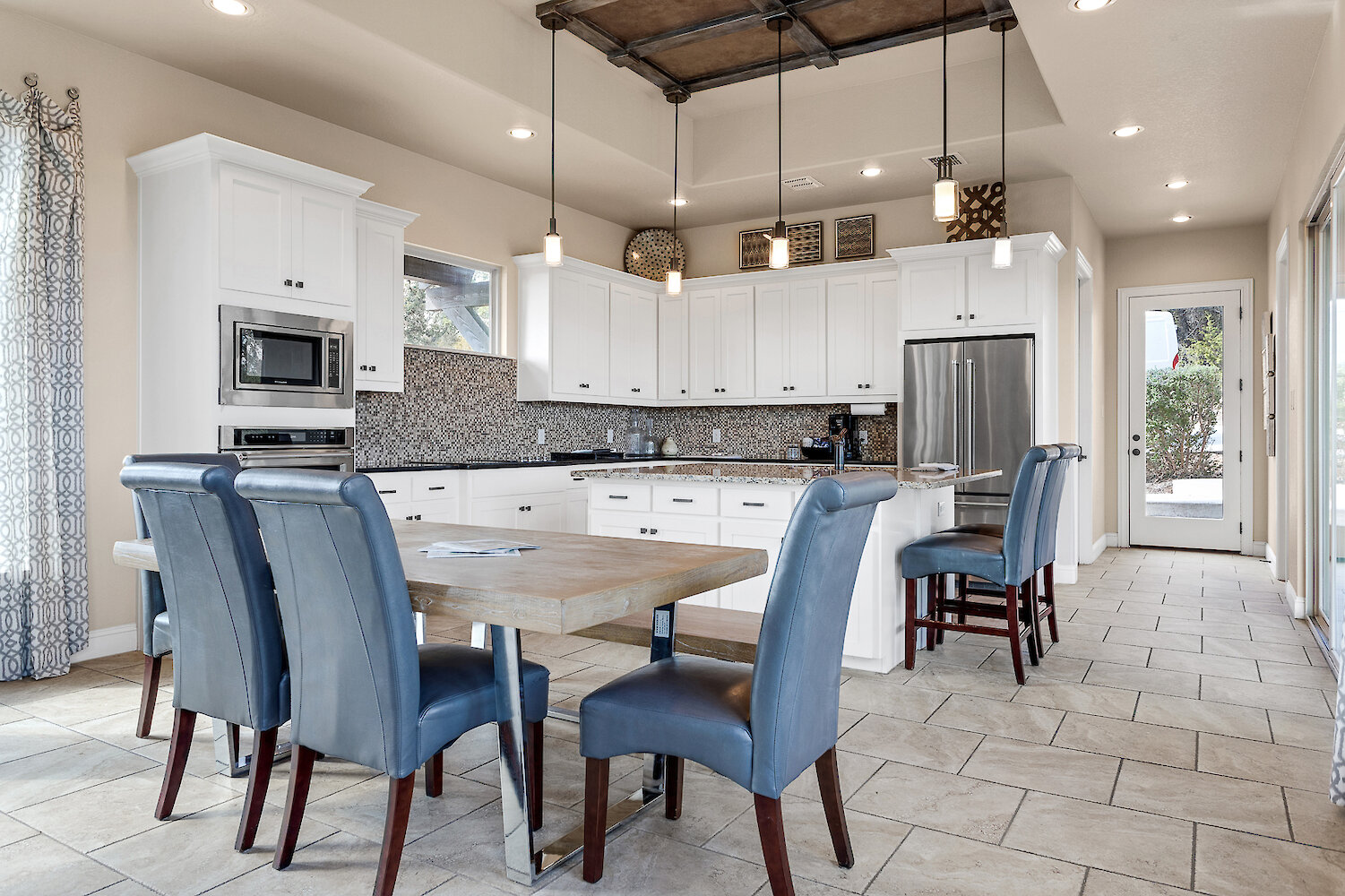 Bright modern kitchen with white cabinets, mosaic tile backsplash, stainless steel appliances, pendant lighting, and a dining table with blue upholstered chairs