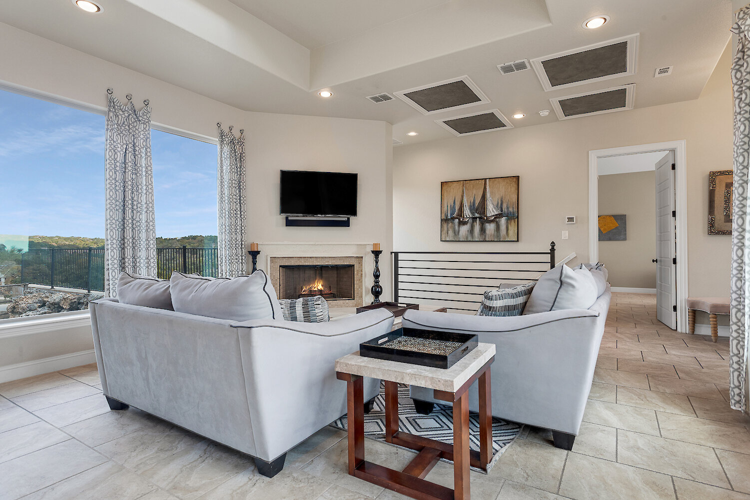 Modern living room with large windows, two gray sofas, tile flooring, a wall-mounted TV above a fireplace, and scenic hill country views.