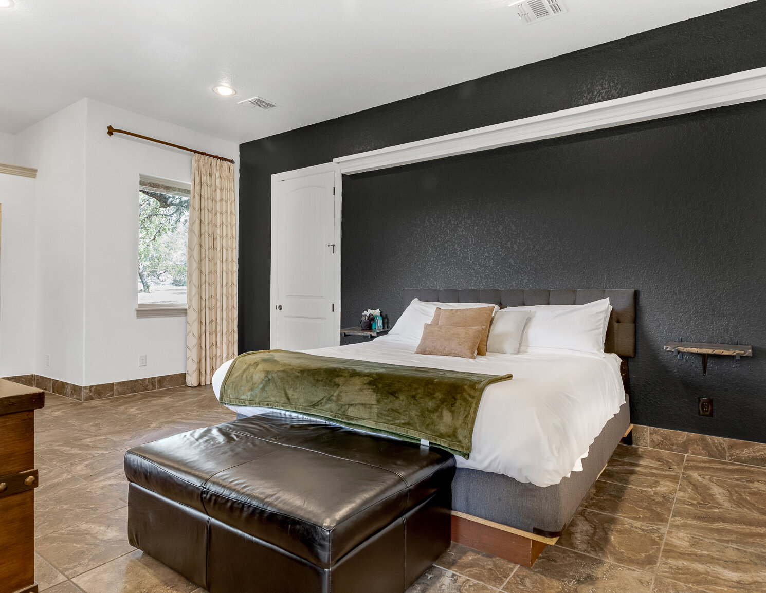 Spacious modern bedroom with a king bed, black accent wall, leather ottoman, tiled floors, and neutral decor.