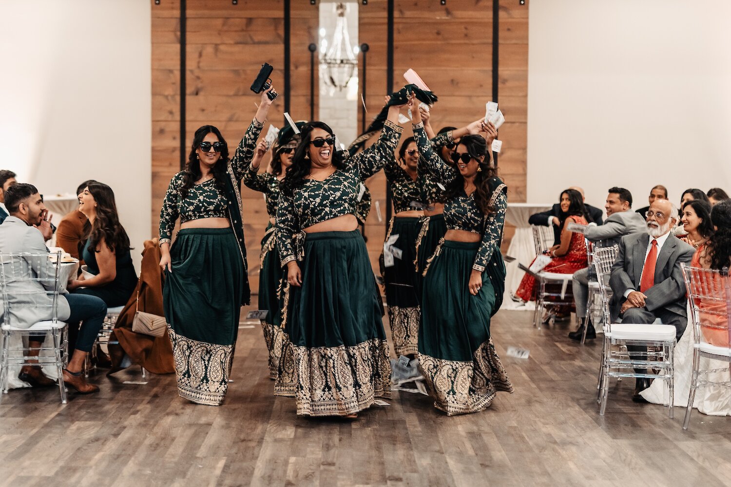 A group of smiling women performing a Bollywood-style dance in matching emerald green lehengas while waving props and money during an Indian wedding sangeet.