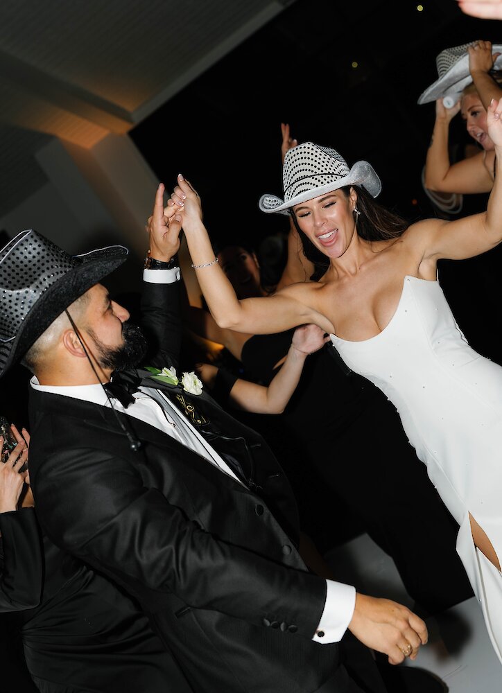 The bride and groom dance enthusiastically at their reception, wearing novelty cowboy hats with the groom in a black tuxedo and the bride in a white gown.