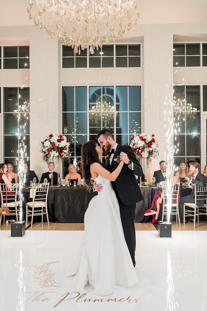 The bride and groom share their first dance on a custom white dance floor with their names, surrounded by indoor cold sparklers and red floral arrangements.
