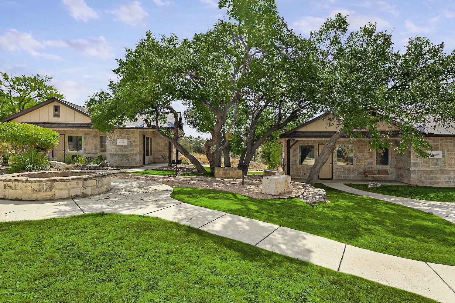 Two exterior stone cottages with dark roofs are set on a grassy lawn with a curved walkway and a stone fire pit in the foreground.