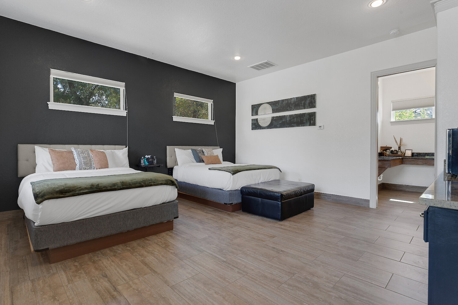 Modern bedroom with two queen beds, dark accent wall, wood-look tile flooring, and adjoining bathroom.