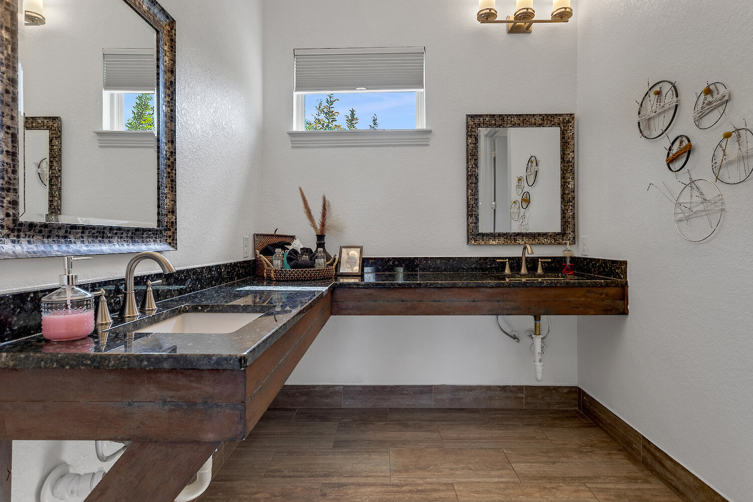 A modern restroom with white walls, dark wood floors, a black marble counter, and decorative mirrors over two sinks.