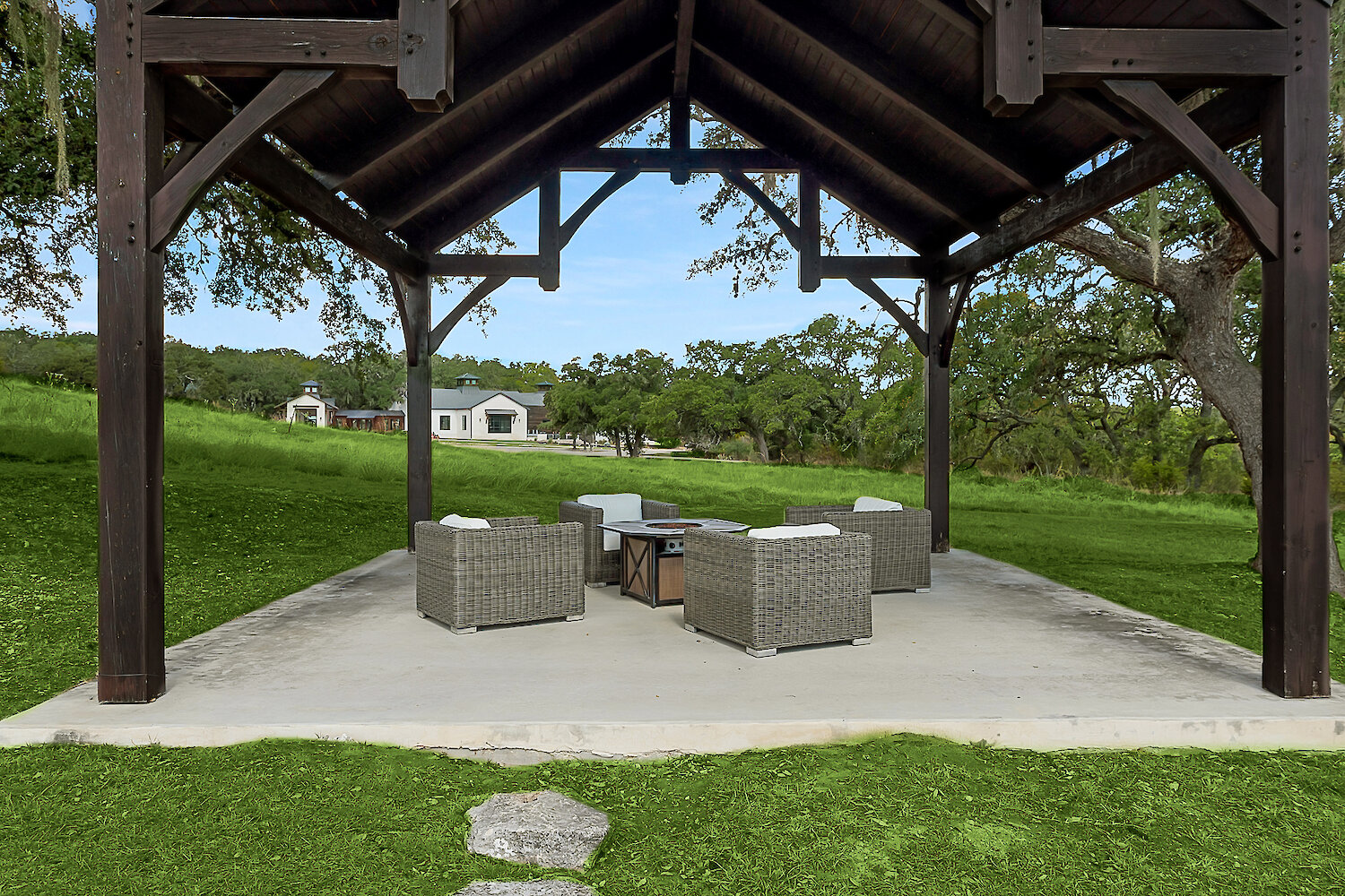 Rustic outdoor pavilion with a dark wood timber roof structure covering a concrete patio. The area features gray wicker outdoor chairs around a metal fire pit table, overlooking a lush green field and distant white farmhouse buildings.
