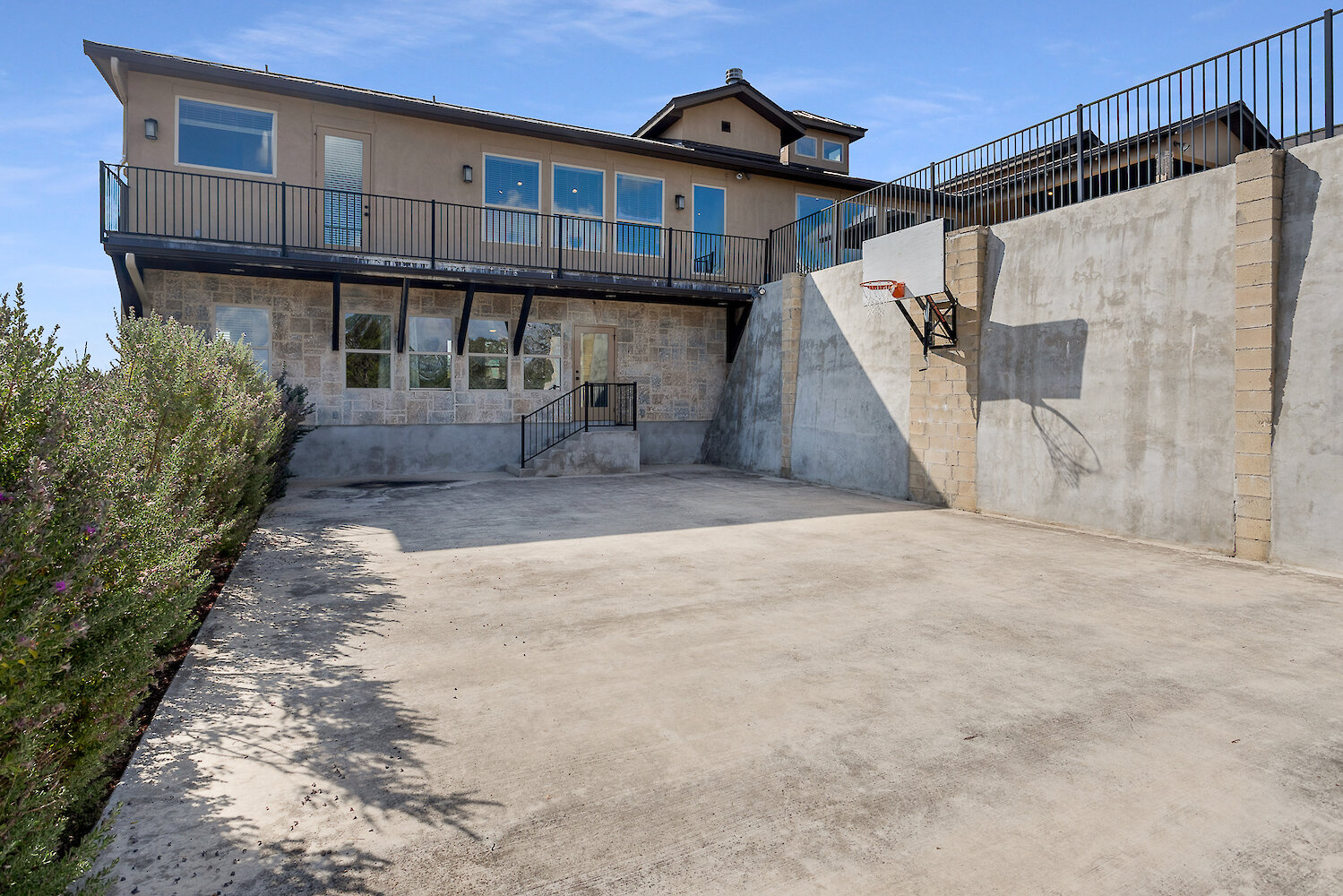 Concrete outdoor basketball court or recreation area built against a large stone retaining wall with an installed basketball hoop, below a multi-story house with a second-floor balcony.