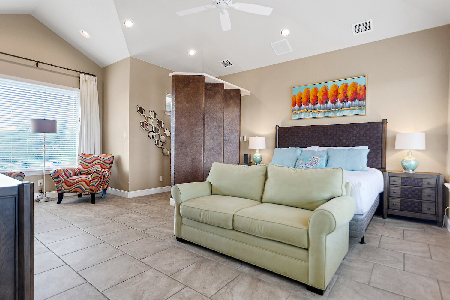 Spacious master bedroom featuring a queen bed with a tufted leather headboard, a light green sleeper sofa, an accent chair, and a decorative room divider on large tile flooring.
