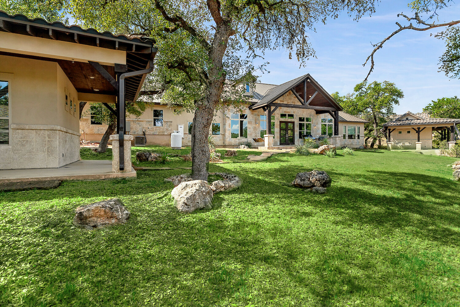Spacious hill country estate with stone exterior, large windows, covered patios, and mature oak trees on a green landscaped lawn.