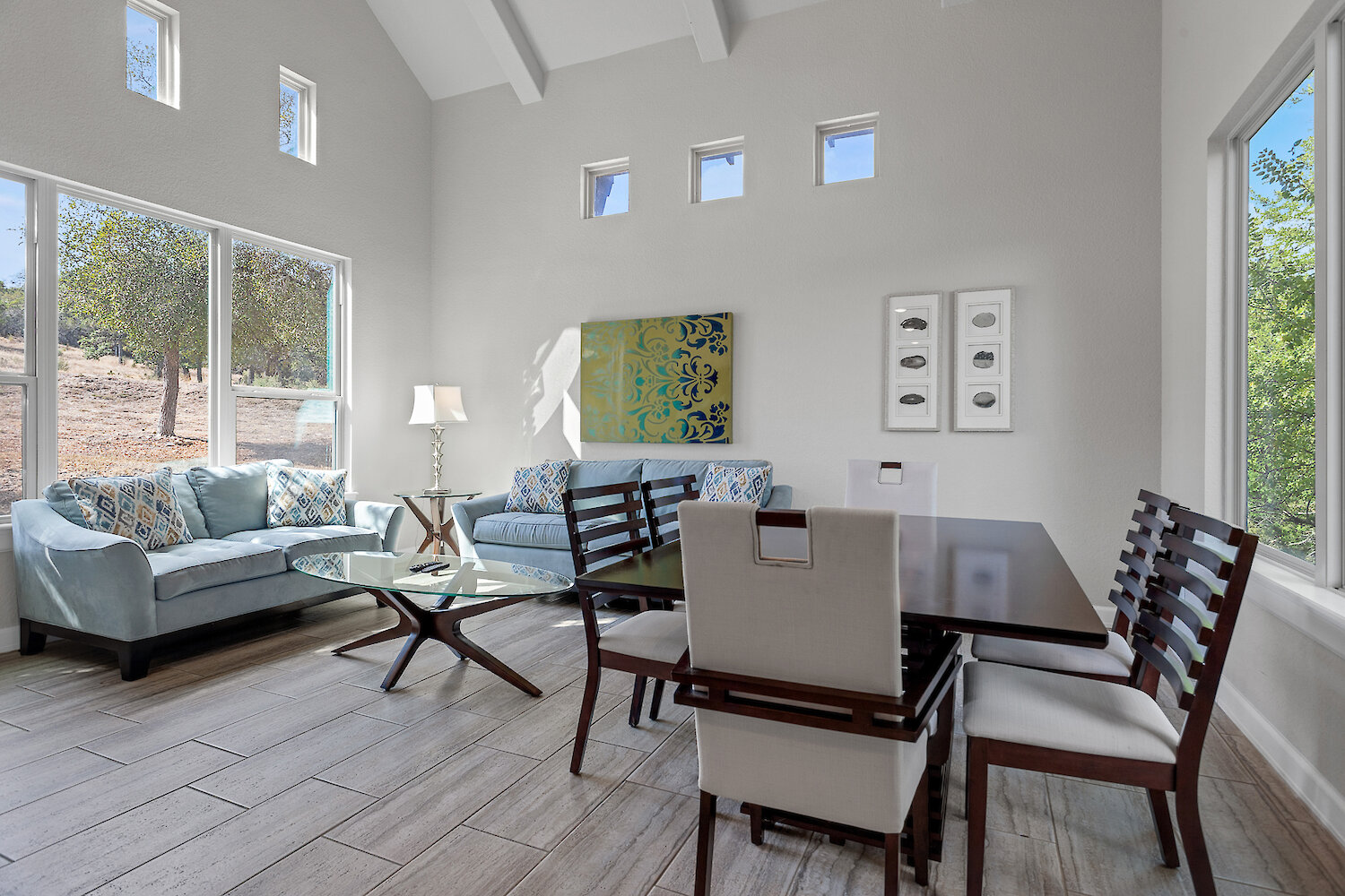 Bright, modern living and dining area with high vaulted ceilings, large windows showcasing a scenic outdoor view, light blue sofas, and a dark wood dining table with upholstered chairs.